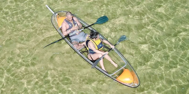 Two people in a clear kayak seen from above, sand visible through the hull